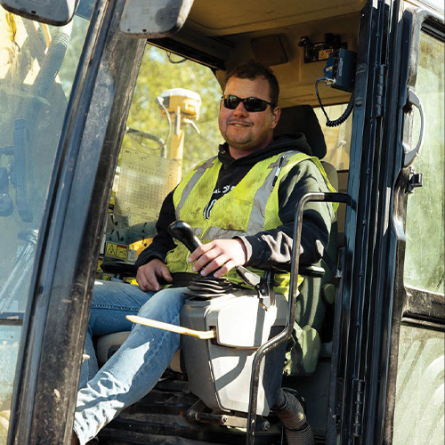 Man in yellow vest and sunglasses smiles in vehicle cab.