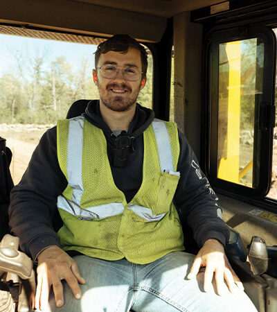 Man in glasses and yellow vest sits in vehicle, trees outside.