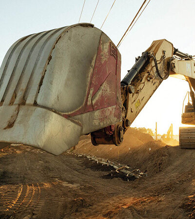 Excavator lifts soil at sunset, power lines above, dirt piles.
