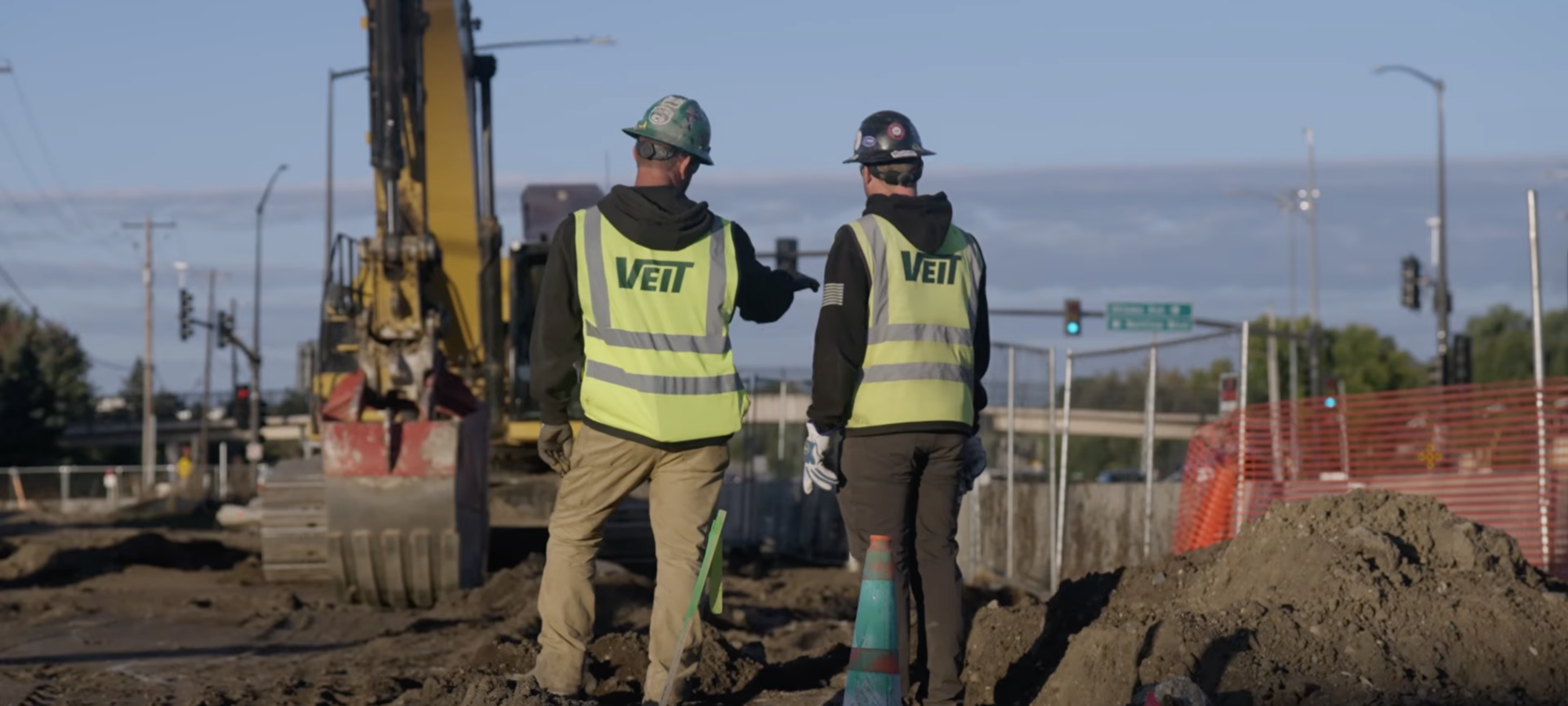 Two workers in safety gear discuss plans by an excavator.