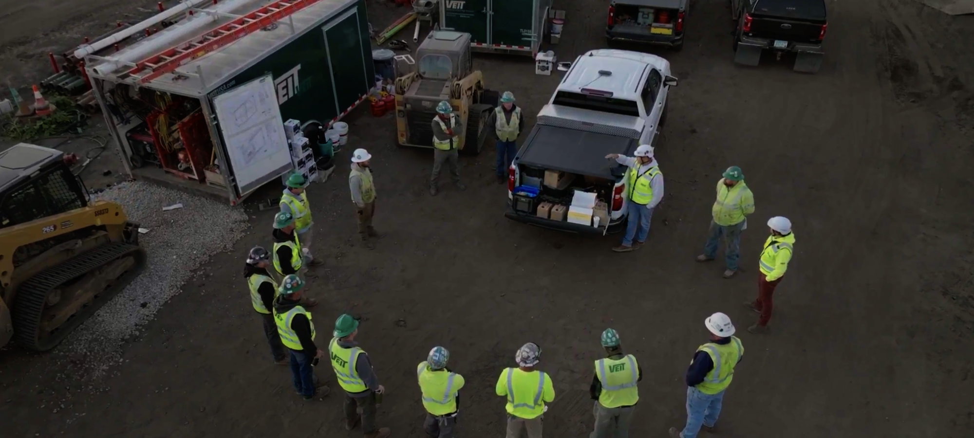 Construction workers in safety gear gather near truck and trailers.