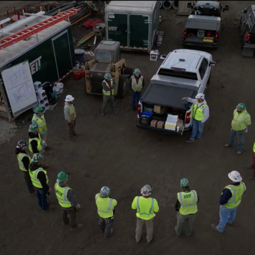 Construction workers in safety gear gather near truck and trailers.