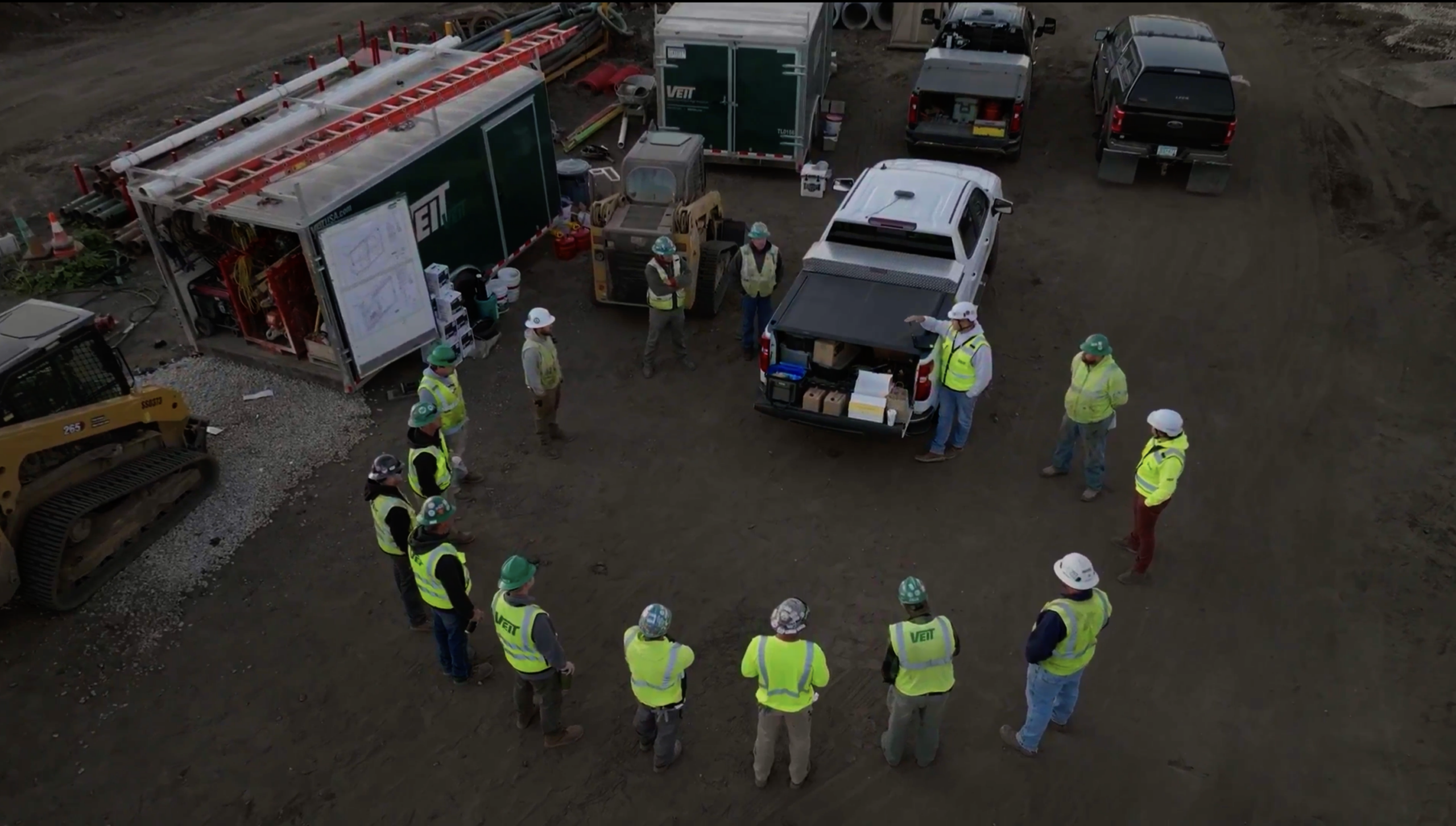 Construction workers in safety gear gather near truck and trailers.