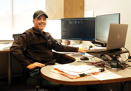 Man in black cap and jacket smiles at desk with monitors.