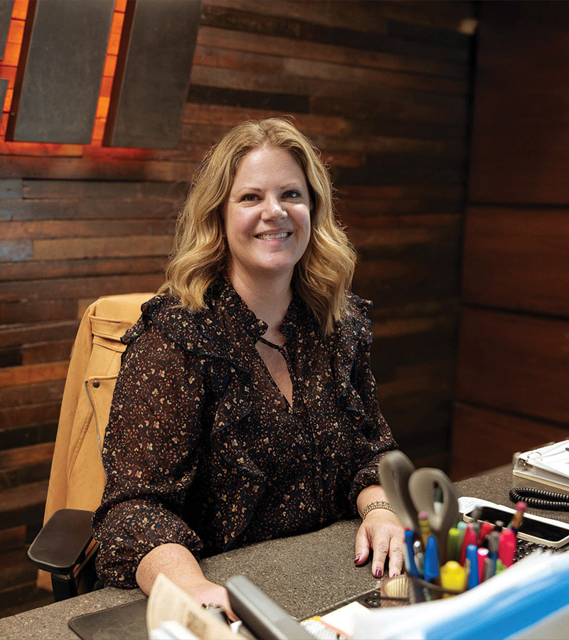 Smiling blonde woman sits at desk with papers; wood-paneled wall.