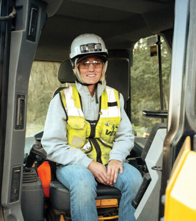 Construction worker in hard hat and vest sits in equipment cab.