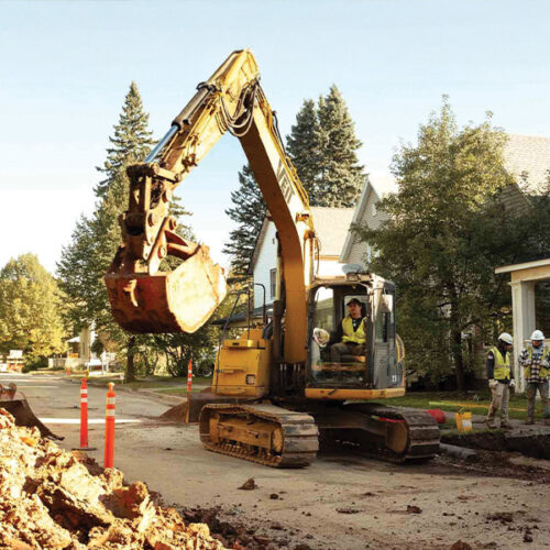 Yellow excavator and workers on street; houses and trees behind.