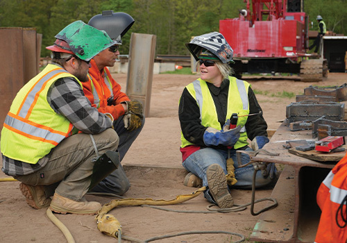 Three construction workers in safety gear talk on a worksite.