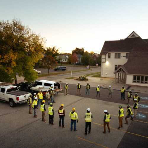 Construction workers in safety gear stand in a parking lot at dawn.