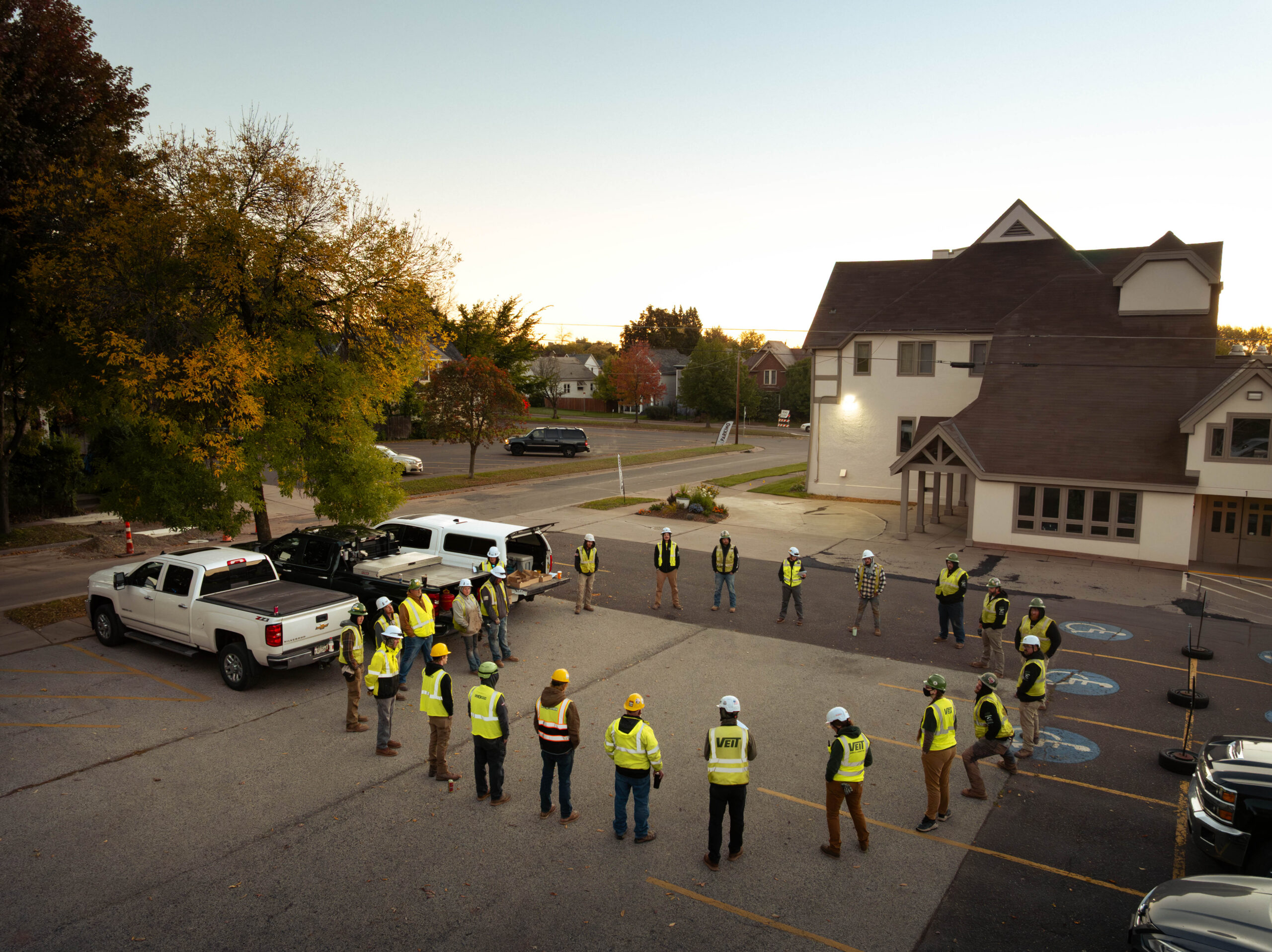 Construction workers in safety gear stand in a parking lot at dawn.