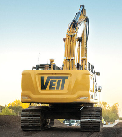 Yellow VEIT excavator on dirt under clear sky; cars behind.