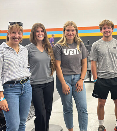 Four young adults smile by a black fence and striped wall.