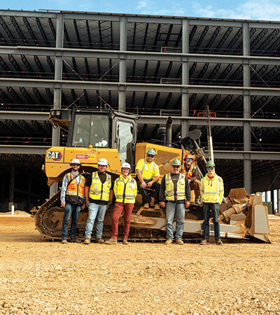 Seven workers in safety gear pose before a bulldozer onsite.