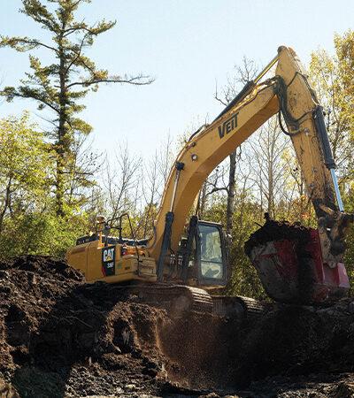 Yellow CAT excavator digs soil in a sunny wooded area.