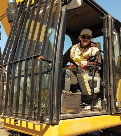 Worker in safety gear sits in yellow excavator cab outdoors.
