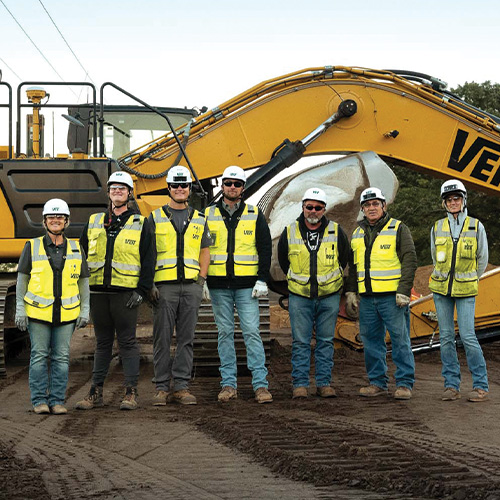 Seven construction workers in safety gear by a yellow excavator.
