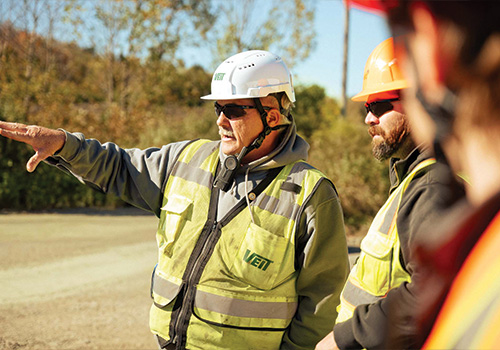 Construction workers in safety gear talk outside near trees.