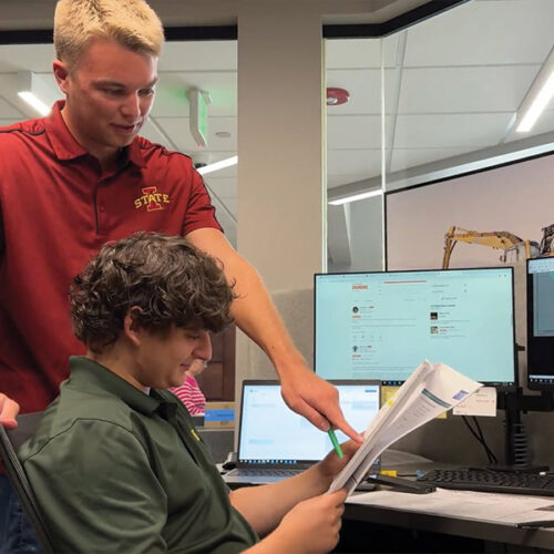 One man stands pointing at papers while another sits at a desk.