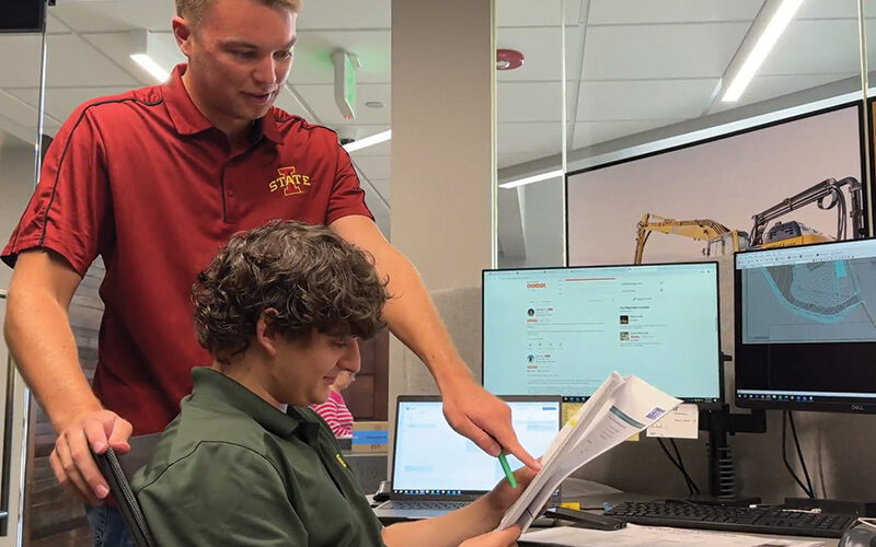One man stands pointing at papers while another sits at a desk.