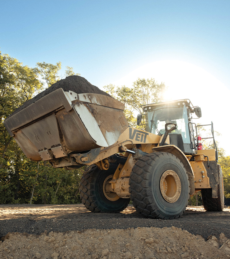 Yellow front loader lifts dirt at sunny, tree-lined construction site.