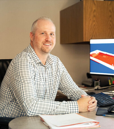 Smiling man at desk with paperwork; 3D model on monitor.