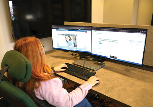 Red-haired person at dual monitors: video call and webpage.