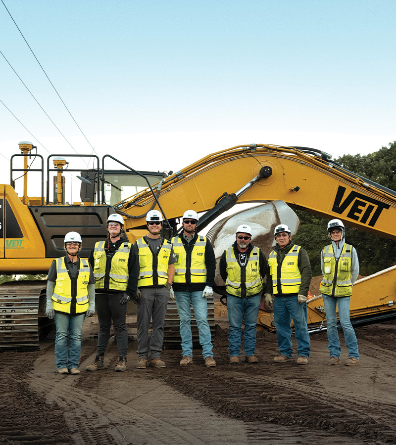 Seven construction workers pose by a yellow excavator on-site.