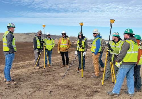 Construction workers in safety gear with surveying equipment listen outside.