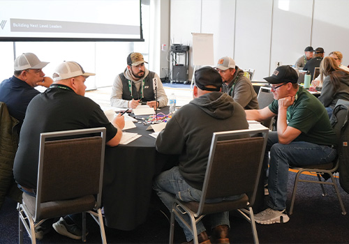 Men discuss at a conference table with papers and presentation screen.