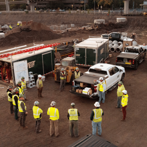 An aerial drone view of a busy construction site: a group of workers in high-visibility vests and hard hats stand in a circle near a mobile trailer, with trucks, equipment, and large pipes, earthworks, and a partially completed overpass in the background.