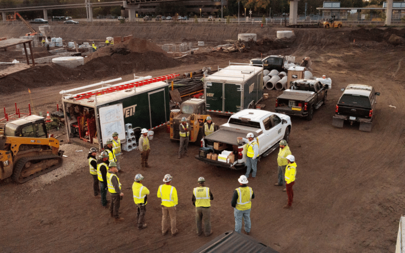 An aerial drone view of a busy construction site: a group of workers in high-visibility vests and hard hats stand in a circle near a mobile trailer, with trucks, equipment, and large pipes, earthworks, and a partially completed overpass in the background.